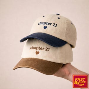May include: Two beige baseball caps, one on top of the other. The top cap has a navy blue brim, the bottom a brown brim. Both caps feature the embroidered text "chapter 21" with a heart symbol, ideal for casual wear.