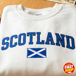 May include: A white crewneck sweatshirt featuring the word SCOTLAND embroidered in bold blue block letters with a dark blue outline. Below the text is an embroidered rectangular flag of Scotland, featuring a white saltire on a blue field.