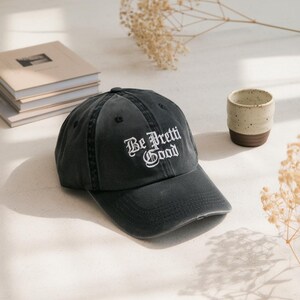 May include: A dark grey baseball cap with the embroidered text "Be Pretti Good" in white script. The cap has a worn appearance and a curved peak. Books and a small ceramic cup are in the background.