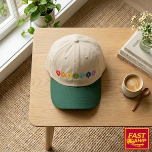 Può includere: Cappellino da baseball beige con visiera verde. Il cappellino presenta una fila di fiori ricamati nei colori dell'arcobaleno. Il cappellino è appoggiato su un tavolo di legno marrone chiaro, accanto a una tazza di caffè e un piccolo vaso di fiori.