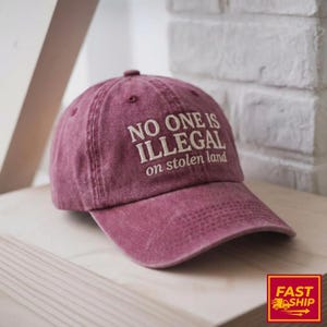 May include: A maroon baseball cap with the embroidered text "NO ONE IS ILLEGAL on stolen land." The cap is made of a soft, worn material and has a curved brim. The cap is sitting on a wooden surface.
