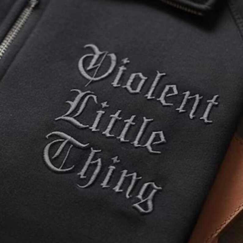 K&ouml;nnte beinhalten: Nahaufnahme eines schwarzen Kapuzenpullovers mit Rei&szlig;verschluss und dem gestickten Text "Violent Little Thing" in einer gotischen Schriftart. Der Rei&szlig;verschluss ist silberfarben, und der Stoff scheint ein weiches Material zu sein.