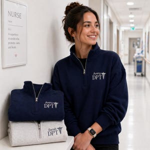 May include: Navy blue quarter-zip sweatshirts with the text "Avery DPT" and a medical symbol. A light gray quarter-zip sweatshirt with the text "Sarah DPT" and a medical symbol is also visible. A framed print reads "NURSE" and defines the word.