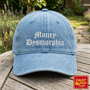May include: Light blue denim baseball cap with the text "Money Dysmorphia" embroidered in white. The cap has a curved brim and an adjustable strap. The hat is sitting on a wooden surface.