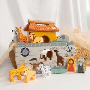 Könnte beinhalten: Ein Holzspielzeug der Arche Noah mit bunten Tierfiguren. Die Arche ist in Gelb, Grau und Weiß bemalt, mit einer Holzrampe. Enthält eine Giraffe, einen Löwen, ein Zebra und andere Tiere. Die Arche trägt den Namen "Noah".