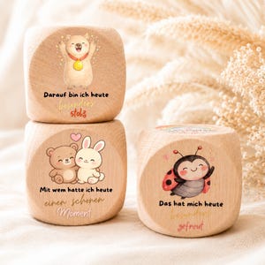 Könnte beinhalten: Drei helle Holzblöcke mit aufgedruckten Illustrationen und deutschem Text. Ein Block zeigt einen Bären mit Goldmedaille, ein weiterer einen Bären und einen Hasen beim Kuscheln und der dritte einen Marienkäfer. Der Text enthält Sätze wie Darauf bin ich heute besonders stolz, Mit wem hatte ich heute einen schönen Moment und Das hat mich heute besonders gefreut.