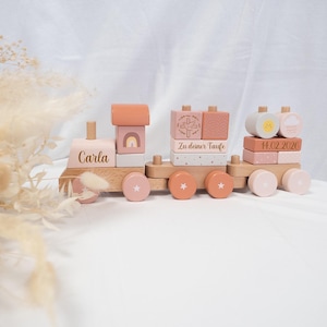 Könnte beinhalten: Ein Holzspielzeugzug mit pastellrosa, pfirsichfarbenen und naturbelassenen Holzblöcken. Der Zug zeigt einen Regenbogen, ein Kreuz und den Namen "Carla". Die Räder sind rosa und pfirsichfarben mit Sternen. Der Text "14.02.2026" ist ebenfalls sichtbar.