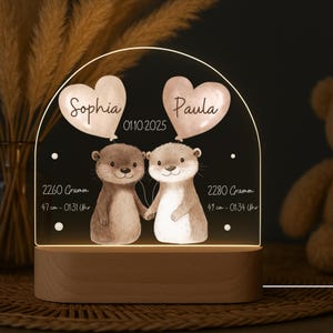 Könnte beinhalten: Personalisiertes Nachtlicht mit zwei Cartoon-Ottern, die Händchen halten. Das Licht zeigt die Namen "Sophia" und "Paula" in herzförmigen Ballons sowie Geburtsdaten: 2260 Gramm, 47 cm, und 2280 Gramm, 49 cm. Auf einem Holzsockel.