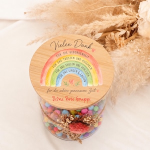 Könnte beinhalten: Ein klares Glasgefäß gefüllt mit bunten kleinen Kugeln, verschlossen mit einem runden Bambusdeckel, der eine Aquarell-Regenbogenillustration und deutschen Text wie Vielen Dank, Für die Geborgenheit und Deine Rote Gruppe zeigt.
