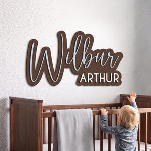May include: A wall-mounted wooden name sign featuring the name Wilbur in a large, dark brown cursive script with a light blue outline, layered over a dark brown backing with the name ARTHUR in white block letters underneath the script.