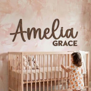 Może przedstawiać: Drewniany napis na ścianę z imieniem Amelia Grace w ciemnobrązowym kroju pisma skryptowego i blokowego. Napis jest zamontowany na ścianie z efektem różowo-białej akwareli, nad jasnym drewnianym łóżeczkiem dziecięcym z pluszowymi zabawkami.