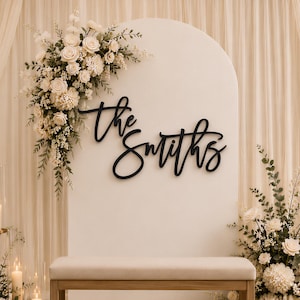 May include: A black script sign reading "the Smiths" mounted on a white arched backdrop. The display includes a wooden bench, cream-coloured floral arrangements with greenery, and several pillar candles in clear glass cylinders on gold stands.