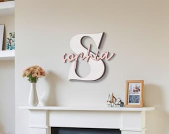 Puede incluir: Decoración mural con una gran letra "S" de madera blanca y el nombre "Sophia" en rosa. La imagen muestra una chimenea, estanterías con libros y objetos decorativos, y un escritorio con una lámpara.
