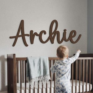 May include: A dark brown wooden wall sign featuring the name Archie in a cursive script font. The letters are connected and mounted horizontally on a wall above a wooden crib. The sign has a smooth, matte finish and a clean, simple design.