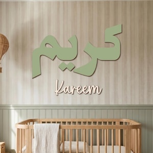 Pode incluir: Um letreiro de parede com o nome Kareem escrito em grande caligrafia árabe verde-sálvia, com o nome Kareem em inglês numa fonte cursiva mais pequena e clara colocada diretamente por baixo do texto árabe numa parede às riscas.
