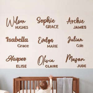 May include: A collection of nine custom wooden name signs mounted on a wall, featuring various first and middle names in a mix of script and block lettering. The names include Wilson Hughes, Sophie Grace, Archie James, Isabella Grace, Evelyn Marie, Julian Cole, Harper Elise, Olivia Claire, and Mason Jude.