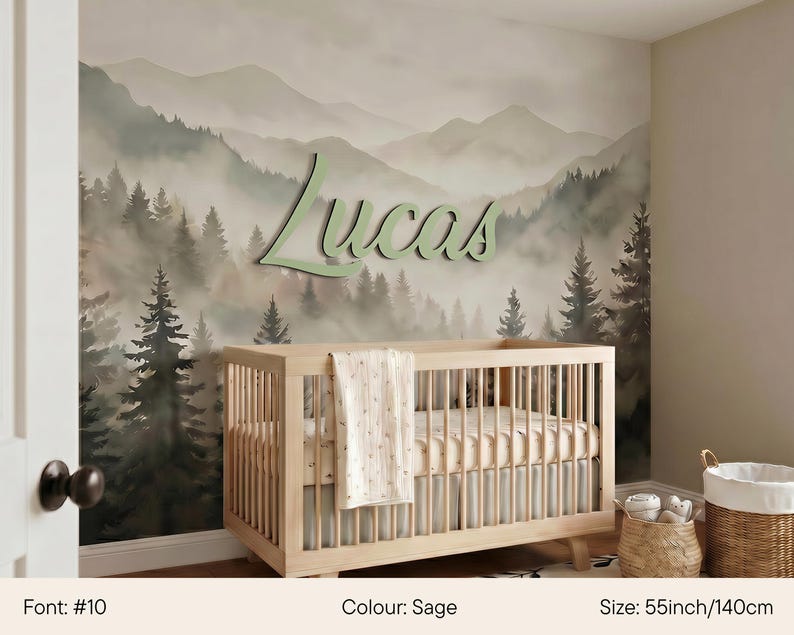 May include: A nursery with a light wood crib and a wall mural depicting a misty forest and mountains. The name "Lucas" is displayed in sage green script. The crib is 55 inches wide.