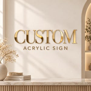 Puede incluir: Un letrero de pared que presenta la palabra CUSTOM en letras grandes y doradas con serifa, con las palabras ACRYLIC SIGN en un texto dorado más pequeño y sin serifa, colocadas directamente debajo de la palabra principal en una pared neutra.