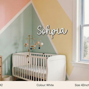 May include: White wooden name "Sophia" in a cursive font, mounted on a wall above a white crib. The room has a pastel color scheme with a geometric design. The name measures 42 inches (107cm) in length.