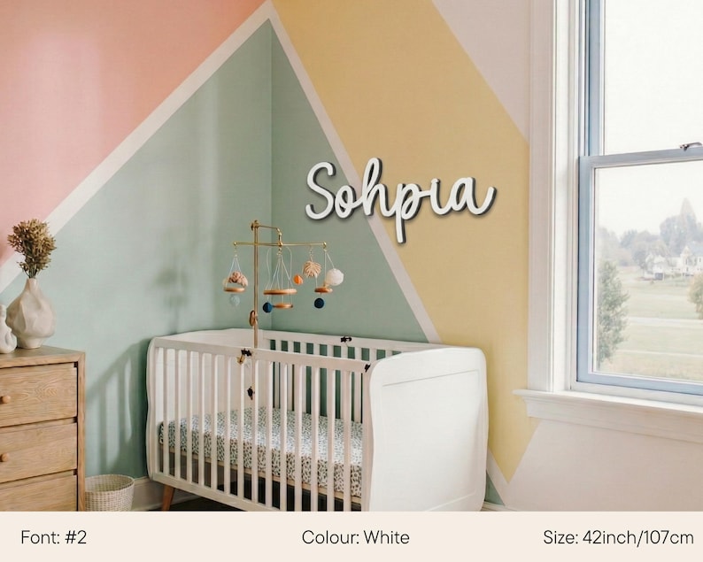 May include: White wooden name "Sophia" in a cursive font, mounted on a wall above a white crib. The room has a pastel color scheme with a geometric design. The name measures 42 inches (107cm) in length.