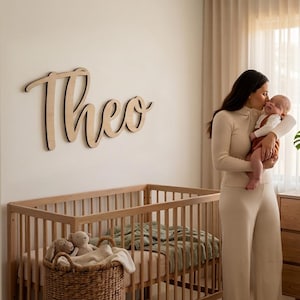 Może przedstawiać: Drewniany znak z napisem "Theo" w kursywie, zamontowany na ścianie. Znak jest z jasnego drewna z czarnym obrysem. W pierwszym planie znajduje się łóżeczko dziecięce i wiklinowy kosz z pluszakami. Osoba trzyma dziecko.