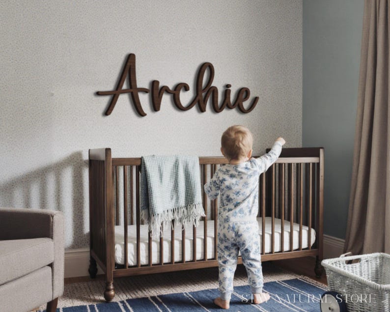 May include: A wooden sign that spells out the name "Archie" is mounted on a wall above a crib. The crib is dark brown and has a blue blanket draped over it. A toddler in a floral onesie stands near the crib.