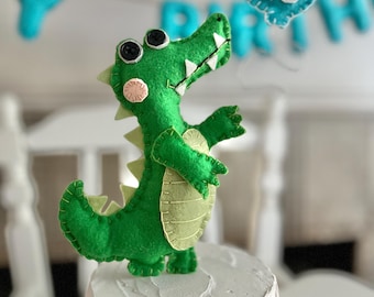 May include: Green felt alligator cake topper with black eyes and a pink nose, holding a blue felt balloon with a white number 2 on it. The alligator is standing on a white cake.