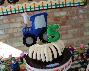 May include: A decorated cake with a blue felt train and a green felt number 5 on top. The cake has white frosting and sits on a decorative stand. Cupcakes with various decorations surround the cake.