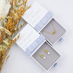 May include: Two white gift boxes with light blue ribbon accents, each containing gold-colored jewelry. One box holds a knot-shaped necklace, the other, knot-shaped earrings with pearl drops. The boxes have the text "I COULDN'T TIE THE KNOT WITHOUT YOU".
