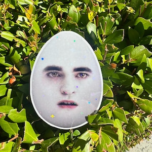 Puede incluir: Un colgante de resina blanca en forma de huevo con un rostro humano con ojos oscuros y labios. El colgante está decorado con pequeñas purpurinas de colores en forma de estrella. El fondo es un primer plano de hojas verdes.