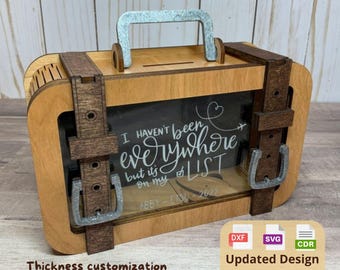 Puede incluir: Una hucha de madera con forma de maleta, con asa y hebillas plateadas. La caja tiene una ventana transparente con el texto "I haven't been everywhere but it's on my LIST". Incluye una ranura para insertar dinero.