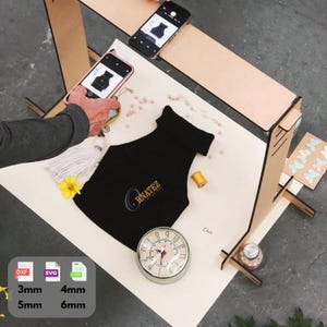 Puede incluir: Un top negro de cuello alto con el logo "CRNATEZ" se muestra sobre una superficie blanca, junto a un reloj vintage, una flor amarilla y una bobina de hilo. Dos smartphones están colocados encima, capturando el top. Una estructura de madera está en el fondo.