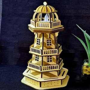 Può includere: Modello di faro in legno con dettagli intagliati. La struttura a più livelli presenta balconi, finestre e una cupola. Il faro è realizzato in legno chiaro e poggia su una base a forma di mattoni. All'interno è visibile una piccola luce.