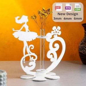 May include: A white laser-cut wooden flower vase with a ballerina silhouette and a heart-shaped design. The vase holds a clear glass tube for flowers and has a flower-shaped base. The text "New Design" is visible.