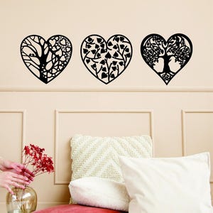 May include: Three black heart-shaped wall decorations with tree and leaf designs. The hearts are mounted on a light beige wall above a bed with white pillows and a red cushion. A vase of red flowers is on the left.