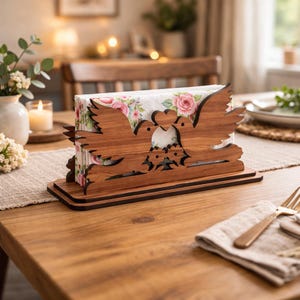 May include: Wooden napkin holder featuring a dove design with a heart between the birds. The holder is made of brown wood and holds floral-patterned napkins. The holder sits on a wooden table with a neutral-colored runner.