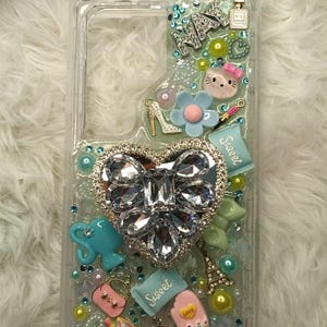 May include: A clear phone case with a glittery, light green background. The case is decorated with various charms, including a large heart-shaped rhinestone, a small Eiffel Tower, a cupcake, and a small ice cream cone. The text "Sweet" appears on the case multiple times.