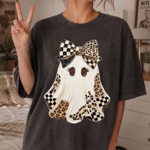 May include: A charcoal gray t-shirt featuring a central graphic of a white ghost wearing a large bow. The ghost design incorporates black and white checkered patterns and brown leopard print accents, with dark brown eyes and long eyelashes.