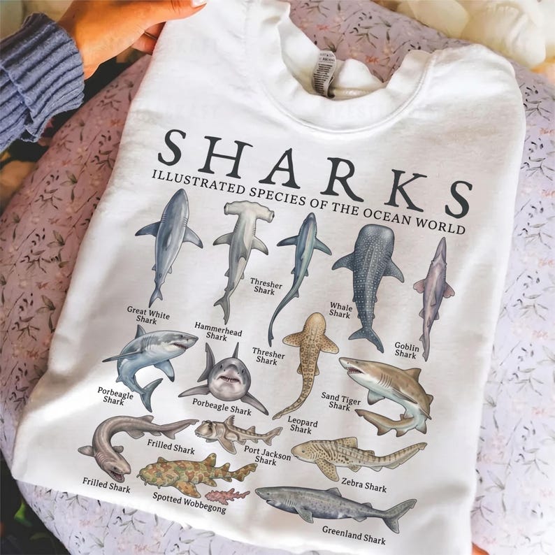 May include: A white t-shirt featuring the text SHARKS ILLUSTRATED SPECIES OF THE OCEAN WORLD and detailed illustrations of various shark species including the Hammerhead, Thresher, Whale, Goblin, Porbeagle, Sand Tiger, Leopard, Frilled, and Greenland shark.