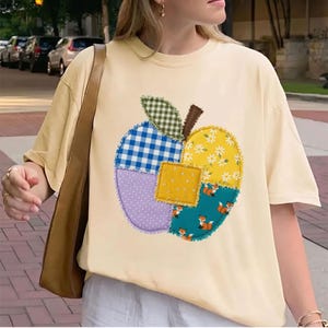 以下が含まれることがあります： 淡い黄色の半袖Tシャツ。様々な布のパッチワークで作られた大きなリンゴの形のグラフィックが特徴です。パッチには、緑と青のギンガムチェック、黄色い花柄、紫の水玉模様、小さなキツネがプリントされたティール色の生地が使われています。