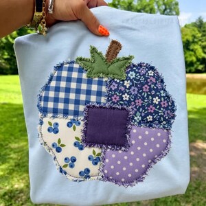 May include: A light blue t-shirt featuring a large, central pumpkin-shaped applique. The pumpkin is constructed from various fabric patches, including blue gingham, floral, blueberry print, and purple polka dot patterns with frayed edges.