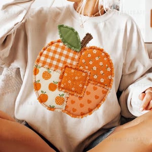 Könnte beinhalten: Ein cremefarbenes Langarm-Sweatshirt mit einer großen, zentralen Applikation in Kürbisform. Der Kürbis besteht aus verschiedenen orangefarbenen und weißen Stoffstücken mit Mustern aus Vichy-Karos, kleinen Blumen, Herzen und kleinen Äpfeln.