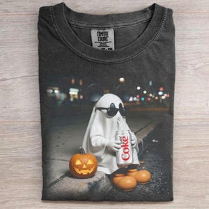 Könnte beinhalten: Ein anthrazitfarbenes T-Shirt mit Rundhalsausschnitt und einem Aufdruck eines weißen Geistes, der eine schwarze Sonnenbrille und Mausohren trägt und eine silberne Dose mit der Aufschrift Coke hält. Daneben steht ein orangefarbener Kürbis.