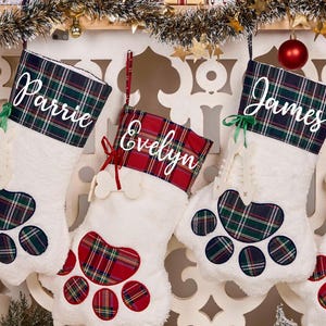 May include: Four white Christmas stockings shaped like paw prints, each with a plaid cuff and a name: Parrie, Evelyn, James, and Noah. The stockings are decorated with red and green plaid paw pads.
