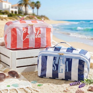 May include: Two striped cosmetic bags with handles sit on a beach. One is coral and white with "Camila" embroidered on it. The other is blue and white with "SEA" embroidered. Sunglasses and beach accessories are in the foreground.