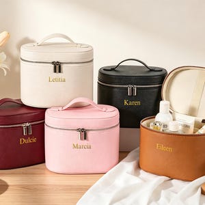May include: A collection of personalized cosmetic bags in various colors, including white, black, pink, burgundy, and brown. Each bag has a top handle, a zipper closure, and a name printed on the front. One bag is open, revealing beauty products.