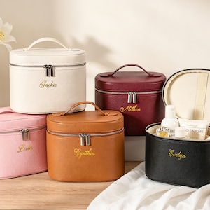 May include: Five personalized cosmetic bags in various colors: white, pink, brown, burgundy, and black. Each bag has a handle, a zipper, and a name embroidered in gold. One bag is open, revealing beauty products.