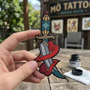 May include: A colorful sticker featuring a dagger piercing a heart. The dagger has a teal handle and a silver blade, wrapped by a red heart. The sticker is held in a hand, with a tattoo shop sign in the background.