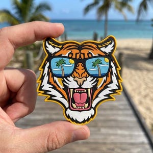 May include: A colorful tiger-shaped accessory with sunglasses reflecting a beach scene. The tiger has orange, black, white, and yellow details, with a wide-open mouth revealing pink and white teeth. The sunglasses show palm trees and a sunny sky.