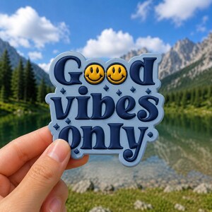 May include: A blue and white sticker with the words "Good Vibes Only" in a playful font. Two yellow smiley faces are above the word "Good". The sticker is held up against a scenic background of mountains and a lake.
